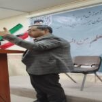 مرد بزرگسالی در حال سخنرانی در مراسمی رسمی با پرچم ایران در دست و تخته‌ی متنی فارسی در پس‌زمینه