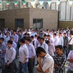 دانش‌آموزان پسر و دختر به لباس فرم مدرسه در حیاط مدرسه جمع‌شده‌اند، ساختمان و تاسیسات مدرسه در پس‌زمینه دیده می‌شود