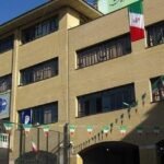 نمای خارجی ساختمان مدرسه با درب و پنجره‌های فلزی و دیوار سفالی زرد و آبی، تزئین با پرچم‌های رنگی