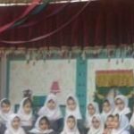 گروهی از دانش‌آموزان نشسته در یک مراسم مدرسه‌ای با پوسترها و تزئینات در پس‌زمینه