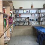کتابخانه مدرسه با قفسه‌های چوبی پر از کتاب، میز مطالعه آبی، و پوسترهای آموزشی روی دیوار