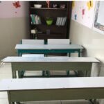 کلاس درس مدرسه‌ای با میزهای سفید، قفسه کتاب‌های درسی، تزئینات دیواری رنگین و آفتاب‌روی