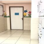 نمای داخلی یک راهرو در مدرسه با درب‌های چوبی، پنجره‌ها، کاشی‌های سفید و تزئینات گل‌های کاغذی روی دیوارها