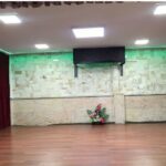سالن مراسم و رویدادهای مدرسه با دیزاین داخلی سبز و قرمز، کف چوبی، و گلدان‌های تزیینی