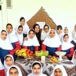 گروهی از دانش‌آموزان دختر در حین یک فعالیت جمعی با میزی پر از میوه‌های تازه و غذاهای رنگارنگ، نشسته به دور هم برای جشن و تعامل اجتماعی