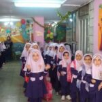 تجمع دانش‌آموزان دختر با روسری سفید و لباس مدرسه‌ای در یک کلاس مزین‌شده با بادکنک‌های رنگی و نقاشی‌های دیواری، حین یک مراسم یا جشن مدرسه‌ای با حضور معلم