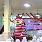 تصویری از یک سالن مراسم و رویداد با صندلی‌های سفید، تزیینات دیواری رنگین، و بنر صورتی رنگ برای یک مراسمی مدرسه‌ای