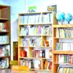 قفسه‌های چوبی پر از کتاب‌های رنگی در کتابخانهِ مدرسه با دیوار فیروزه‌ای و کره‌ی جغرافیایی