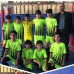 گروه تیمی دانش‌آموزان با پیراهن یکسان سبز و زرد در کنار یک معلم، ایستاده در سالن ورزشی مدرسه برای مراسم یا مسابقه