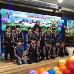 گروه دانش‌آموزان با لباس تکریمی در صحنه‌ای تزئین شده با بالون‌های رنگی و بنر، به مناسبت یک مراسم یا رویداد مدرسه‌ای.