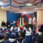 دانش‌آموزان نشسته در حالی که برنامه‌ای با اجرا کنندگان روی صحنه تزیین‌شده با بنرهای رنگی برگزار می‌شود