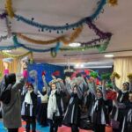 دانش‌آموزان دختر در حال اجرای برنامه رقص جمعی در مراسمی با تزئینات رنگی در سالن مدرسه