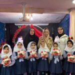 گروهی از دانش‌آموزان دختر با روسری و یونیفرم آبی به همراه معلمان در مراسم تقسیم هدایا در اتاقی با تزئینات قرمز و گل‌های مصنوعی