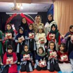 گروهی از دانش‌آموزان در یک مراسم مدرسه‌ای به همراه معلم، هر کدام در حال نگه‌داشتن هدیه یا صنایع‌دستی قرمز رنگ