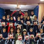 دانش‌آموزان دختر در یک مراسم کتاب یا فعالیت فرهنگی در محیط مزین‌شده با دیوارهای آبی، هر کدام یک بسته‌ی کتاب یا هدیه در دست دارند