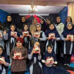 گروهی از دانش‌آموزان دختر مدرسه به لباس فرم در حال نمایش هدایای بسته‌بندی شده در مراسم جشن یا تقدیری درون کلاسی با دیوارهای آبی رنگ