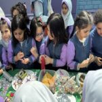 دانش‌آموزان دختر و پسر دبستان در حال شرکت در یک جشنواره غذایی مدرسه، ایستاده اطراف میز سبز پر شده با غذاهای مختلف، لباس فرم آبی و بنفش