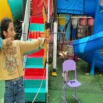 دانش‌آموز در حیاط بازی مدرسه با تجهیزات رنگین شامل لغزنده آبی و سلاید‌های قرمز و فیروزه‌ای