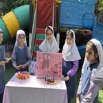 دانش‌آموزان دختر در حیاط مدرسه حول میزی در یک رویداد یا نمایشگاه مدرسه‌ای