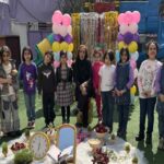 دانش‌آموزان دختر مدرسه در حیاط با بالن‌های صورتی و تزئینات موقت، شرکت در مراسمی یادبود که با گل‌های تازه و ساعت‌های نمادین مزین است
