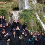 گروهی از دانش‌آموزان دختر با لباس سیاه در یک تور مدرسه‌ای، ایستاده جلوی آبشار طبیعی در کوهستان