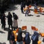 دانش‌آموزان و معلمان در حیاط مدرسه حول صندلی‌های نارنجی جمع‌آوری شده، نشانه‌ی برگزاری یک رویداد یا مراسمی در محوطه خارجی