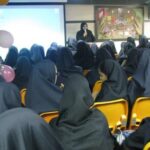 دانش‌آموزان دختری در کلاس درس در حین مراسمی یا رویدادی، نشسته در صندلی‌های زرد، معلمی در جلوی کلاس ایستاده‌است