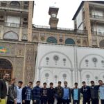 گروهی از دانش‌آموزان و معلمان در مقابل یک بنای تاریخی معماری ایرانی سنتی با تزئینات کاشی‌کاری رنگی و درب‌های قوسی ایستاده‌اند