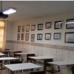 کلاس درس مجهز با میزهای دانش‌آموزی، پنجره روشن با پرده، دیوار‌های تزیین‌شده با نقاشی‌های متعدد در قاب، پروژکتور سقفی و روشنایی تخصصی