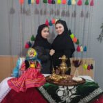 دو دختر دانش‌آموز با پوشش سنتی‌یِ فرهنگی در حین جشنواره‌ی فرهنگی مدرسه، کنار تزئینات رنگین و آثار هنری سنتی