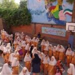 گردهمایی دانش‌آموزان در حیاط مدرسه با پوشش یکدست نارنجی و سفید، پس‌زمینه دیوار آبی با تابلو و پرچم‌های ایران