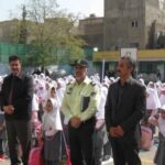 دانش‌آموزانی در لباس یکدست (سفید و صورتی) در صفوف منظم در حیاط مدرسه ایستاده‌اند، معلمان و مسئولین مدرسه در جلو درحال نظارت هستند