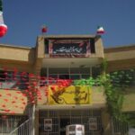 نمای بیرونی ساختمان مدرسه‌ای با معماری سفید و صفحه‌ای، تزئین‌شده با پرچم‌های ایرانی و رنگ‌های قرمز سبز روی دیوار