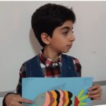 دانش‌آموز پسری که یک کار هنری (ماهی رنگین‌کمان) را نشان می‌دهد و در کلاس درس نشسته است