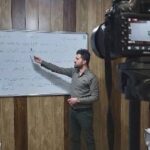 یک مدرس در حال توضیح مطالب روی تخته‌ی سفید پر از متن فارسی، در حالی‌که یک دوربین حرفه‌ای محتوای آموزشی را برای ضبط و پخش در حال ضبط است