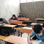 کلاسی با دانش‌آموزانی که در میزهای خشتی نشسته‌اند و درحال کار هستند، دیوار پشتی تزئین‌شده با نقوش بتنی.