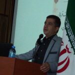 یک سخنران در حال سخنرانی و پومن‌خوانی در جلسه‌ای رسمی با میکروفون، پرچم ایران و نشان تکریم‌الشاعران در پس‌زمینه