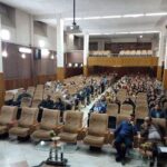 سالن برگزاری مراسم در مدرسه با صندلی‌های متعدد و حضور دانش‌آموزان در حال تجمع