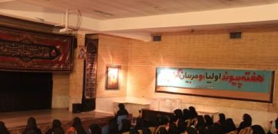 دانش‌آموزان در حال شرکت در مراسم و اجتماع مدرسه‌ای در سالن با صحنه و تابلوهای معلوماتی