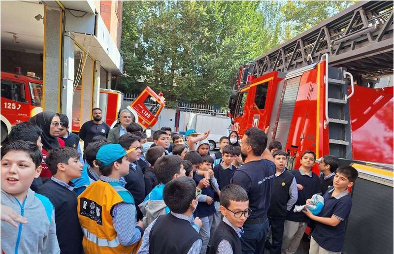 دانش‌آموزان در حال بازدید از ایستگاه آتش‌نشانی و آموزش ایمنی با حضور کارکنان ایمنی و تجهیزات آتش‌نشانی قرمز