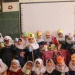 گروهی از دانش‌آموزان دختر با لباس‌های سنتی و رسمی در حال حضور در یک مراسم یا رویداد مدرسه‌ای داخل کلاس درس، در کنار تخته‌سیاه و تزئینات دیواری.
