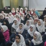 گروه بزرگی از دختران با حجاب سفید نشسته در یک محل داخلی برای شرکت در مراسم یا جلسه‌ای