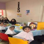 گروهی از دانش‌آموزان دختر در حال شرکت در یک فعالیت اجتماعی گروهی که دور میزی نشسته‌اند و کتاب‌ها و وسایل روی میز قرار دارند.