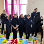 دانش‌آموزان دختر در حال شرکت در یک فعالیت تعاملی گروهی دور تشک رنگین با حروف فارسی، معلمان در حال نظارت