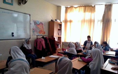 دانش‌آموزان در محیط کلاس درس به دور میز‌های تحریر نشسته‌اند. اتاق دارای تخته هوشمند روی دیوار، پنجره‌های پردار، قفسه‌های کتاب، و محیط نمونه کلاس درسی معمول مدرسه است. دانش‌آموزان زن در حال مشارکت در درس
