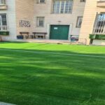 حیاط مدرسه با چمن مصنوعی سبز و صندلی‌های قرمز در کنار ساختمان