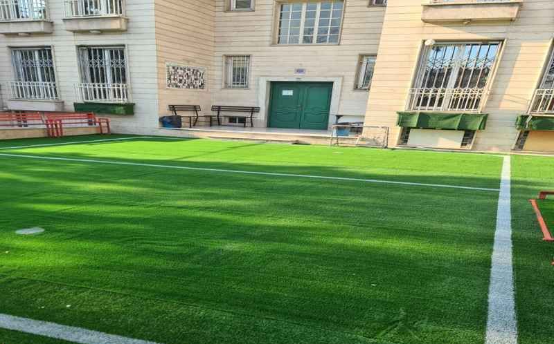 حیاط مدرسه با چمن مصنوعی سبز و صندلی‌های قرمز در کنار ساختمان