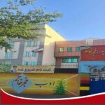 نمای بیرونی ساختمان مدرسه با دیوارهای خاکستری و قرمز، تابلوی معلومات دائمی روی دیوار، و پرچم ایران