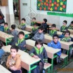 دانش‌آموزان پایه‌ی ابتدایی در کلاس درس مجهز به میز و صندلی‌های سبز رنگ، دیوار تزیین‌شده با پوسترهای رنگی