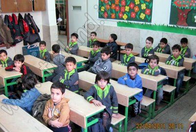 دانش‌آموزان پایه‌ی ابتدایی در کلاس درس مجهز به میز و صندلی‌های سبز رنگ، دیوار تزیین‌شده با پوسترهای رنگی